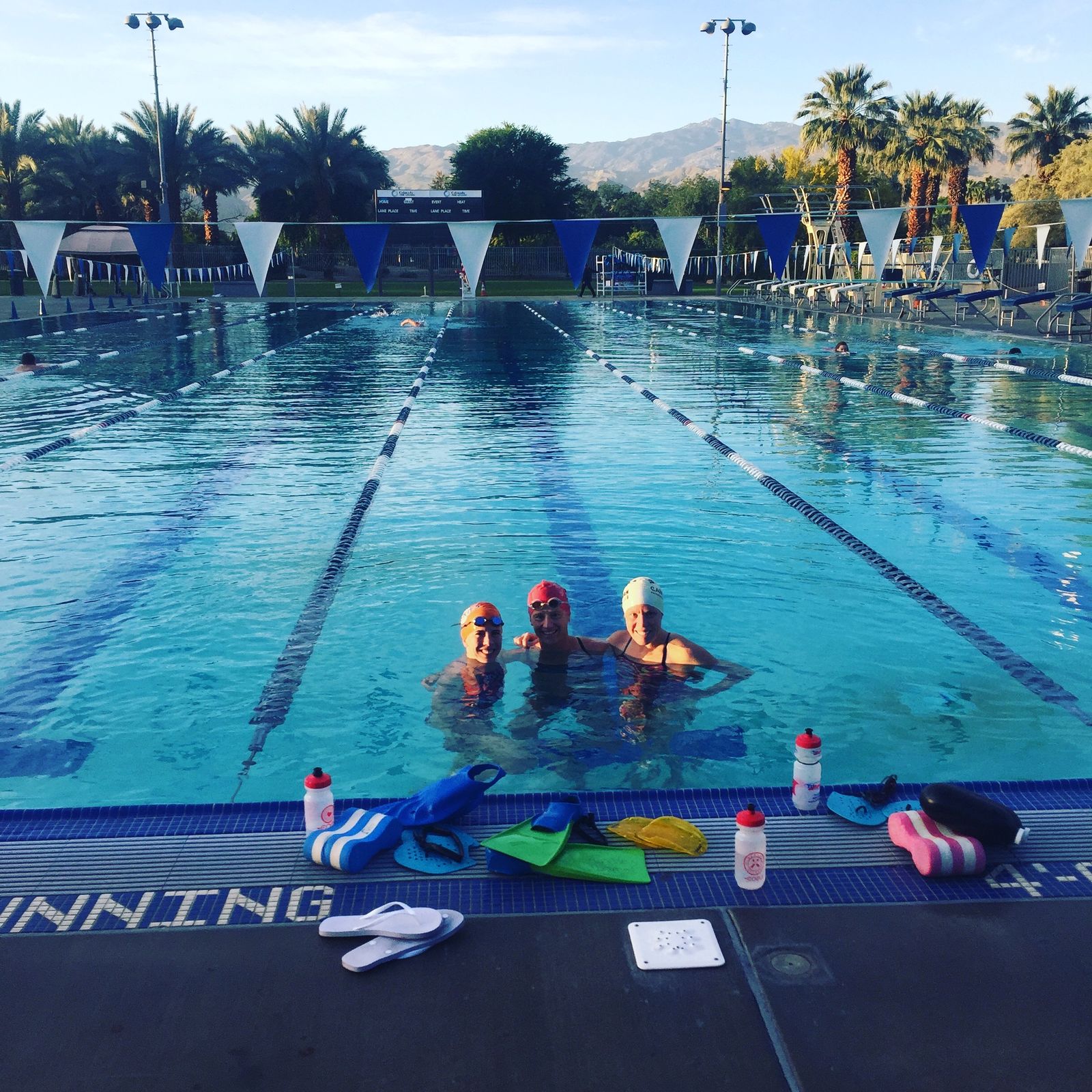 Three swimmers in an outdoor pool with kickboards, fins and paddles on the deck